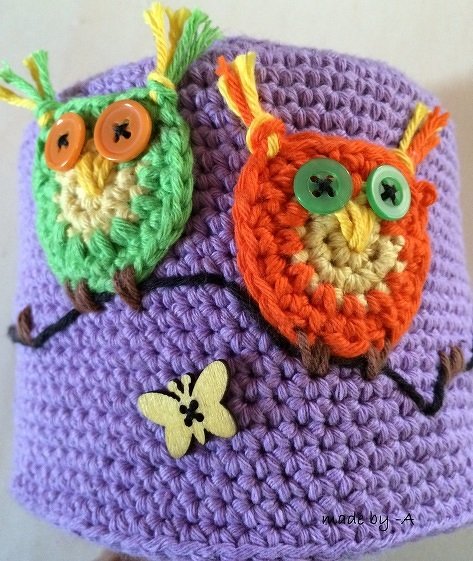Nahaufnahme eines lilafarbenen gehäkelten Überzugs mit zwei applizierten Eulen (grün, orange) mit Knopfaugen auf einem dunklen Ast und einem kleinen gelben Holzschmetterling.