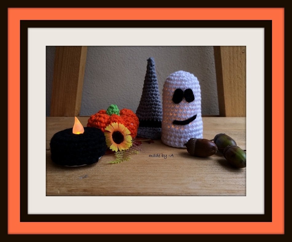 Gehäkelter Kürbis, gehäkelter Geist und gehäkelter Hexenhut auf Holztisch, arrangiert mit Kerze, Sonnenblume und Eicheln.