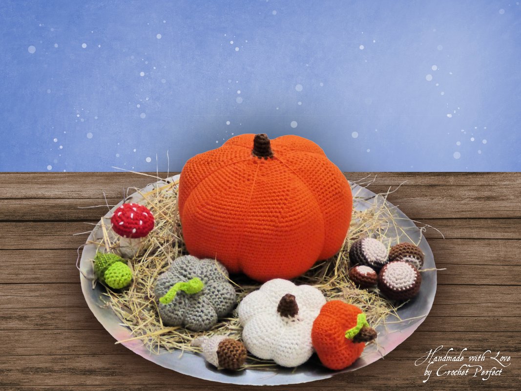 Tablett mit gehäkelter Herbstdeko: großer oranger Kürbis, kleinere Kürbisse, roter Fliegenpilz, Eicheln und Kastanien auf Stroh vor Holzuntergrund.