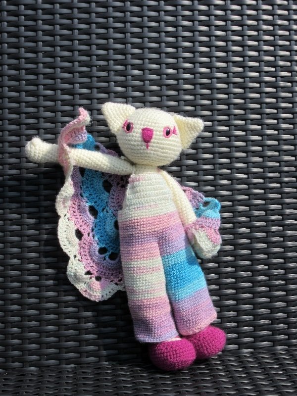 Gehäkelte Amigurumi-Katze in Pastellstreifen mit Umhang auf geflochtenem Untergrund