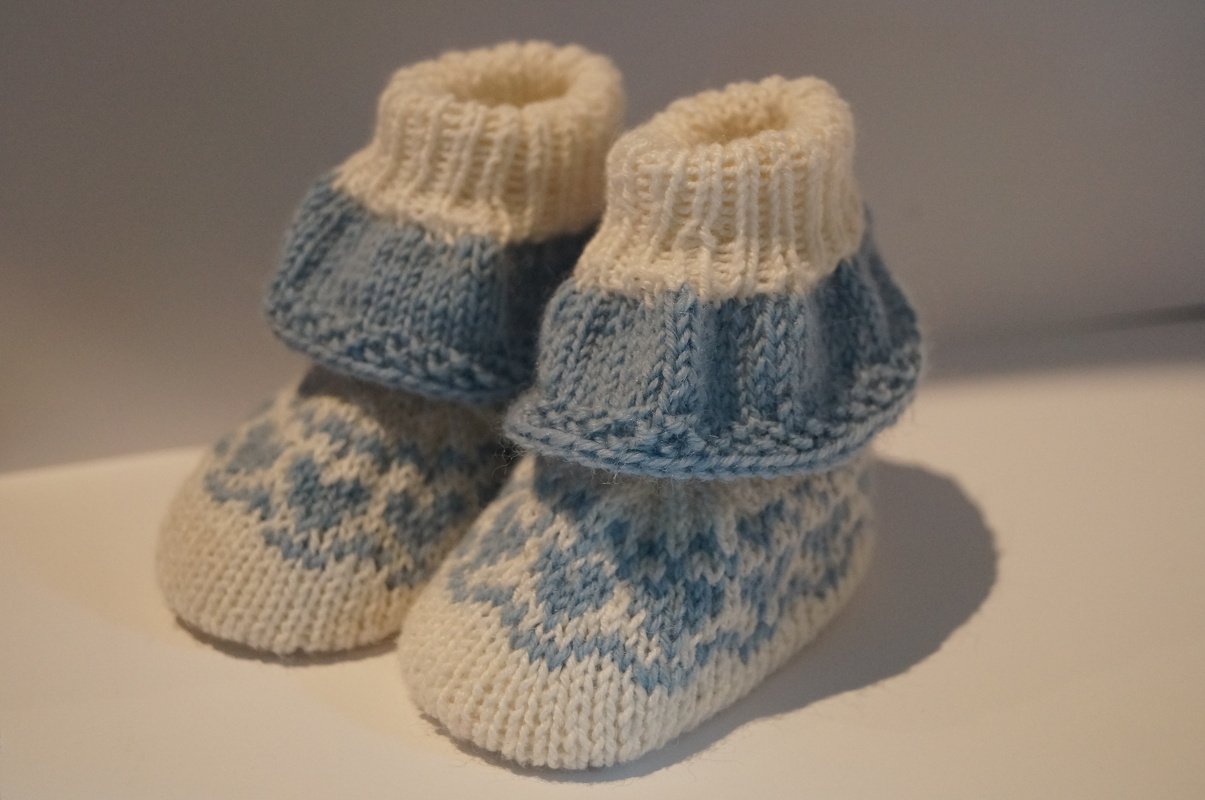 Ein Paar gestrickte Babyschuhe in Hellblau und Cremeweiß mit geometrischem Fair‑Isle‑Muster und geripptem Schaft.