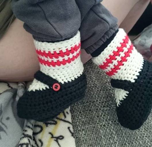 Gehäkelte Babyschuhe in Schwarz mit weiß-roten Streifen und rotem Knopf am Riemen, getragen von einem Kind.
