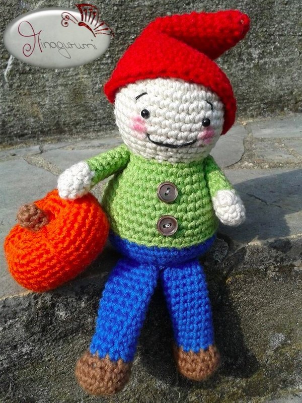 Gehäkelte Wichtelfigur mit roter Mütze, grünem Oberteil, blauen Hosen, braunen Füßen und einem orangefarbenen Kürbis