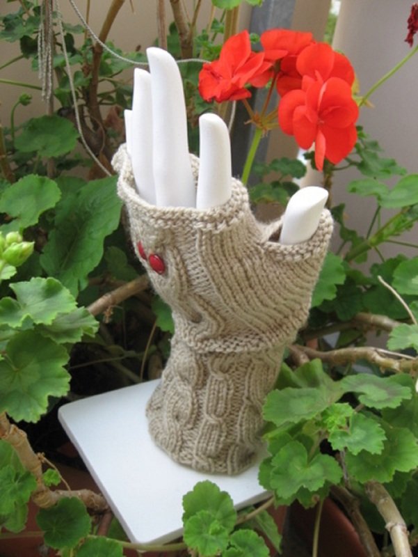 Beiger, fingerloser Strickhandschuh mit Zopfmuster auf weißer Handpuppe, umgeben von grünen Pflanzen und roten Blüten.