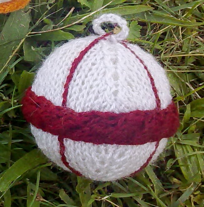 Weiße gestrickte Weihnachtskugel mit dunkelrotem Band und Streifen auf Gras.