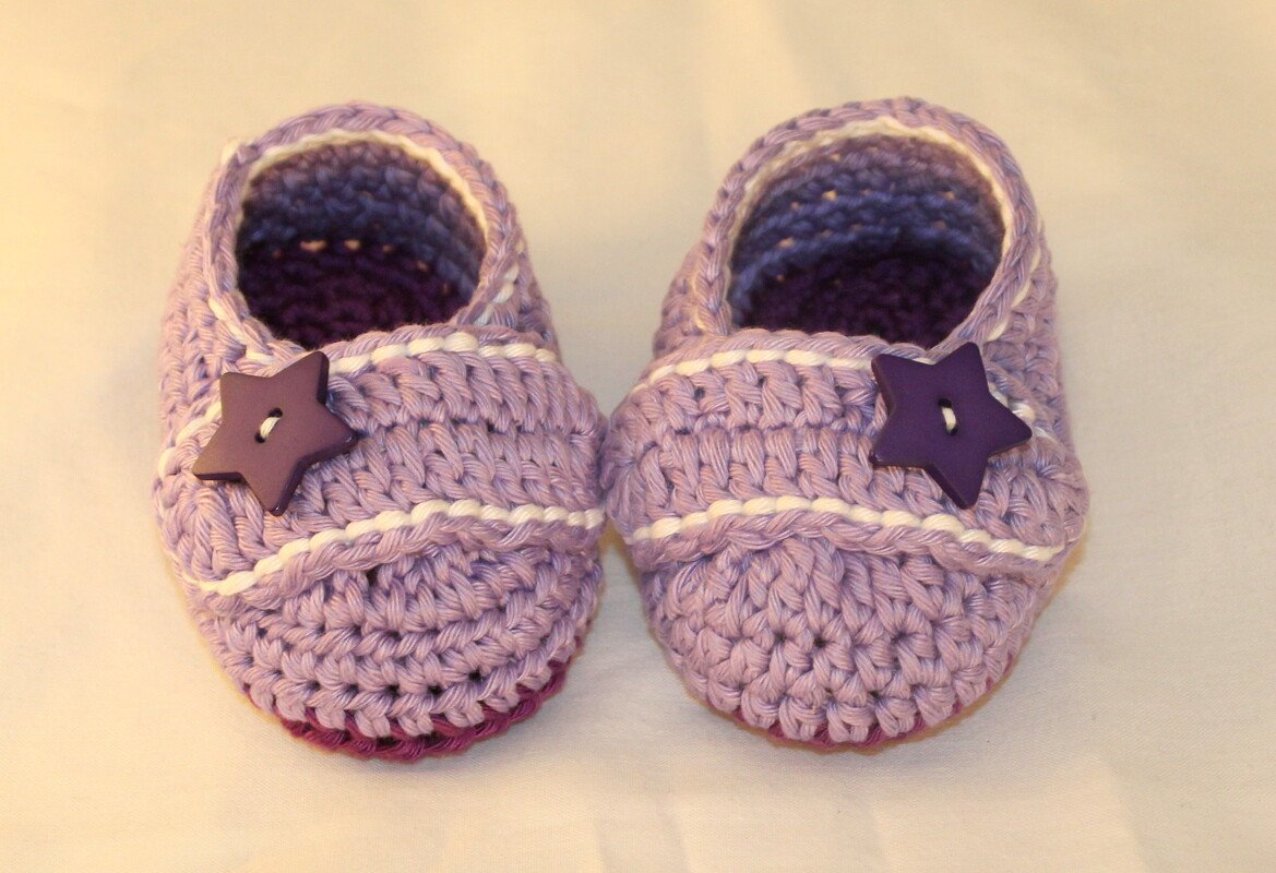 Ein Paar lila gehäkelte Babyschuhe mit weißen Streifen und dunklen Sternknöpfen von oben.