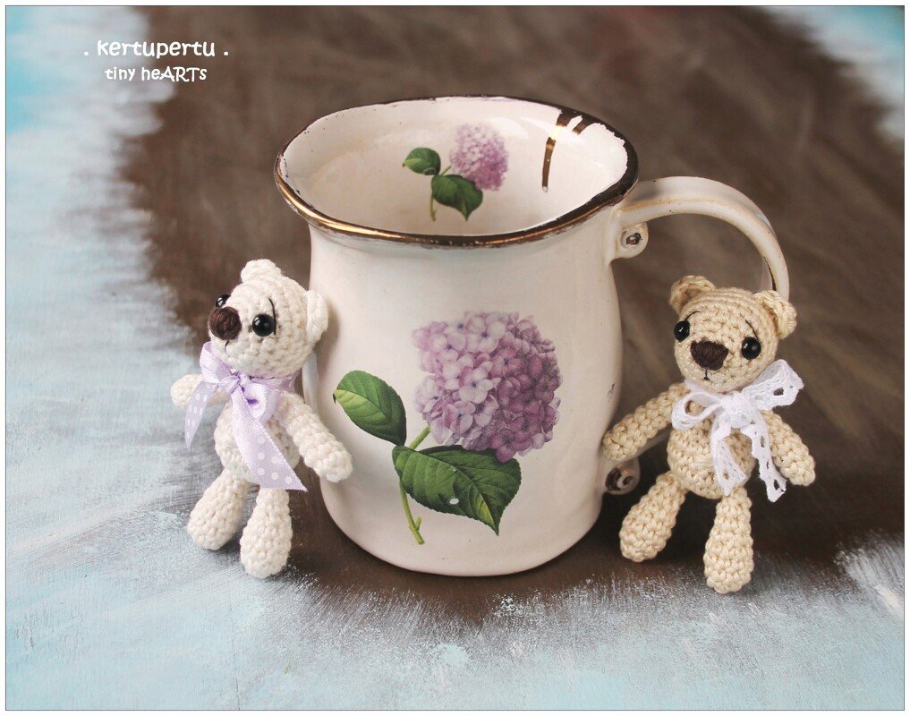Zwei kleine gehäkelte Amigurumi-Teddybären in Creme und Beige neben einer Tasse mit Hortensienmotiv.
