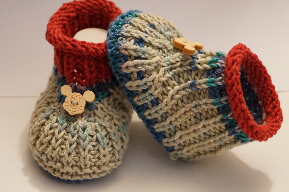 Nahaufnahme gestrickter Babyschuhe mit sichtbarer Maschenstruktur, rotem Bündchen und Holzknopf mit kindlichem Motiv