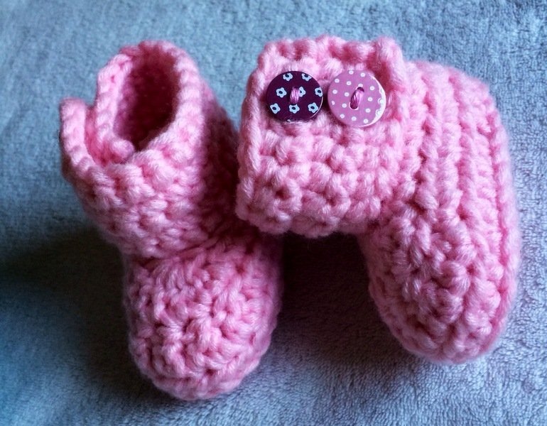 Zwei rosa gehäkelte Babyschuhe mit Umschlag und zwei Zierknöpfen auf hellem Stoff