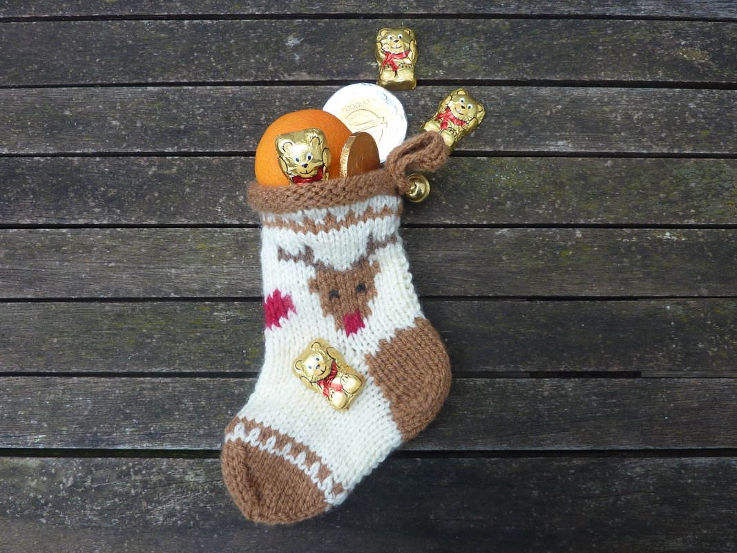 Gestrickter Weihnachtsstrumpf in Weiß und Braun mit Rentier-Motiv, gefüllt mit Orange und Schokolade auf Holzoberfläche.
