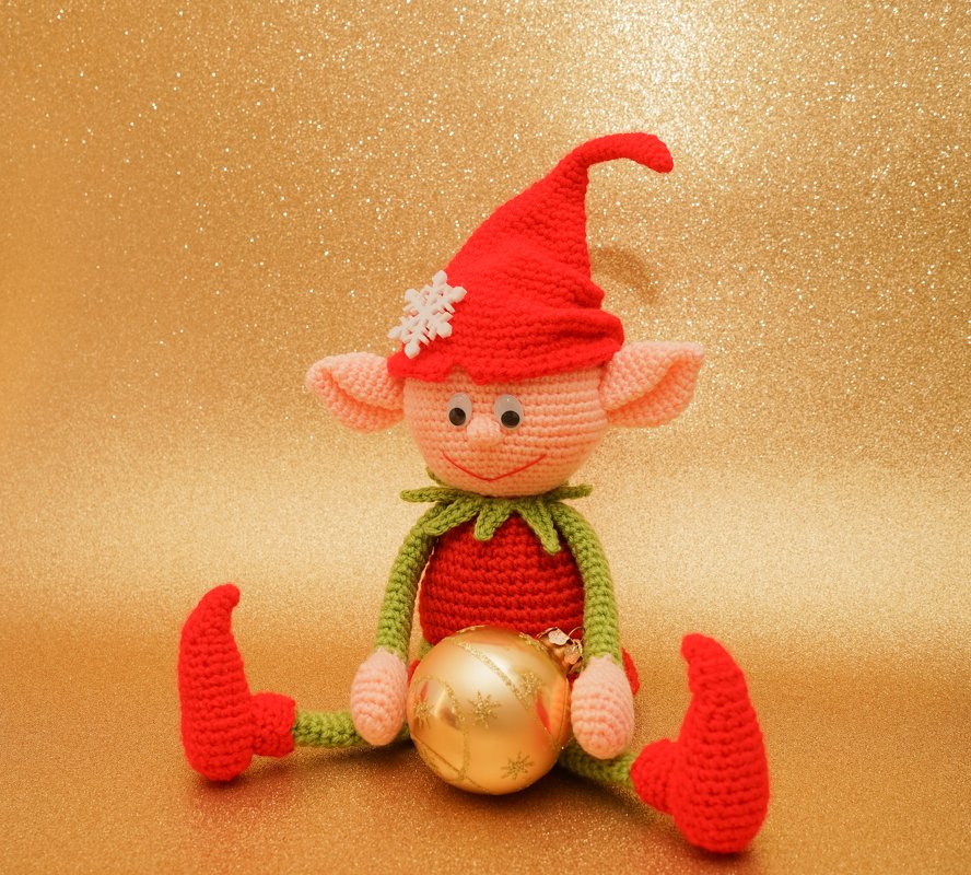 Amigurumi-Wichtel in Rot und Grün, sitzend und eine goldene Weihnachtskugel haltend vor goldglitzerndem Hintergrund.
