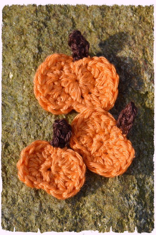Drei orange gehäkelte Kürbisse mit braunen Stielen auf moosigem Untergrund.