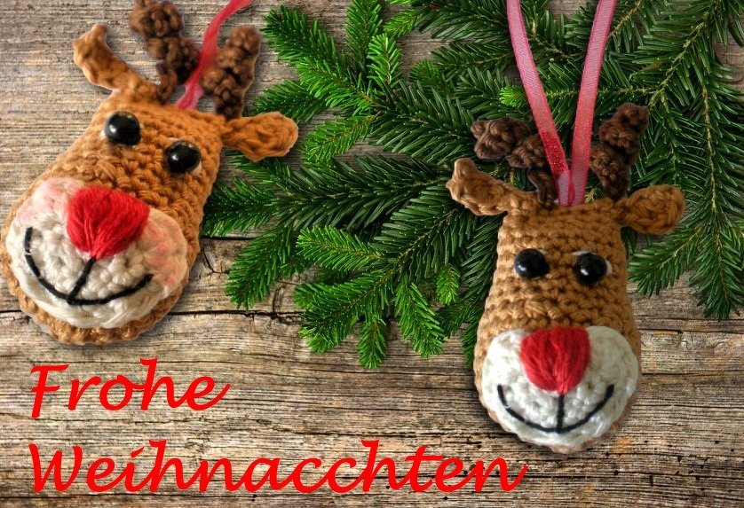 Zwei handgehäkelte Rentierköpfe mit roten Nasen, schwarzen Knopfaugen und braunen Geweihen auf Holz mit Tannenzweigen und rosa Bändern.