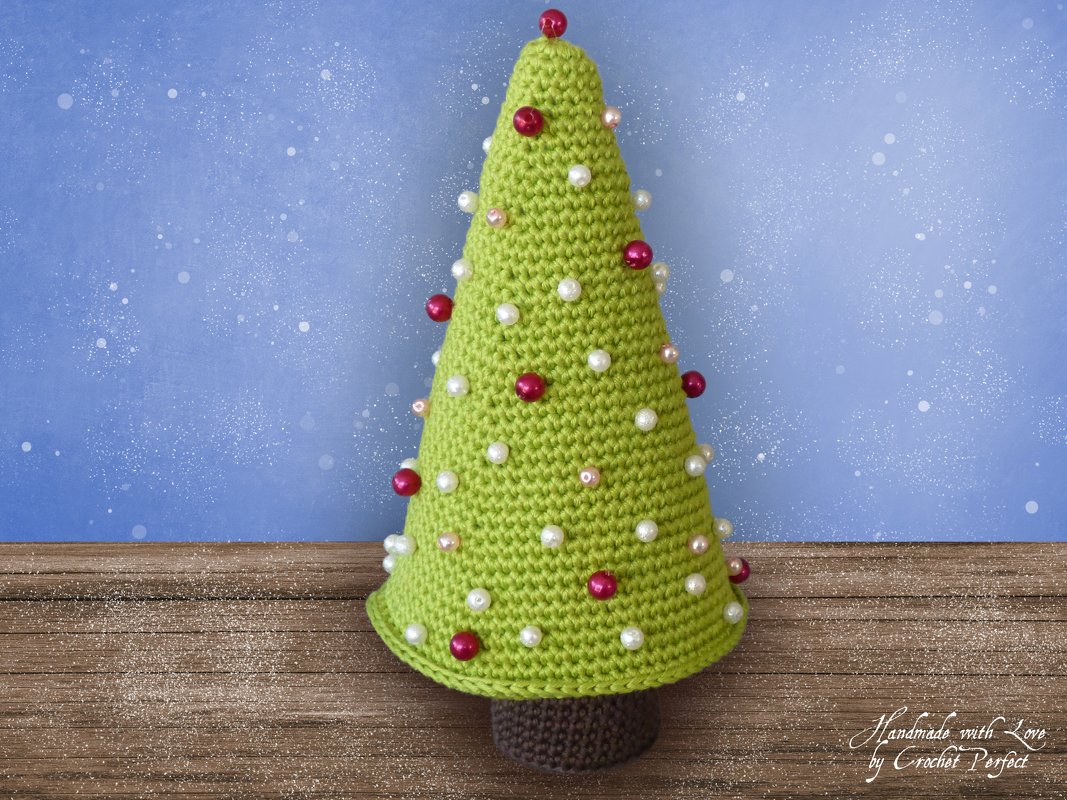 Gehäkelter grüner Tannenbaum mit roten und weißen Perlen auf braunem Sockel vor blauem Hintergrund