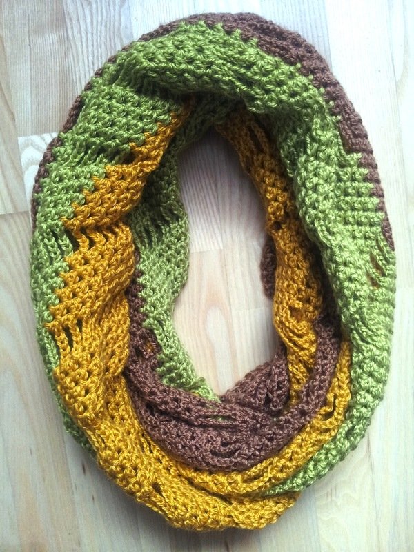 Crocheted infinity scarf in olive green, golden yellow, and brown with a braided texture on a wooden background.