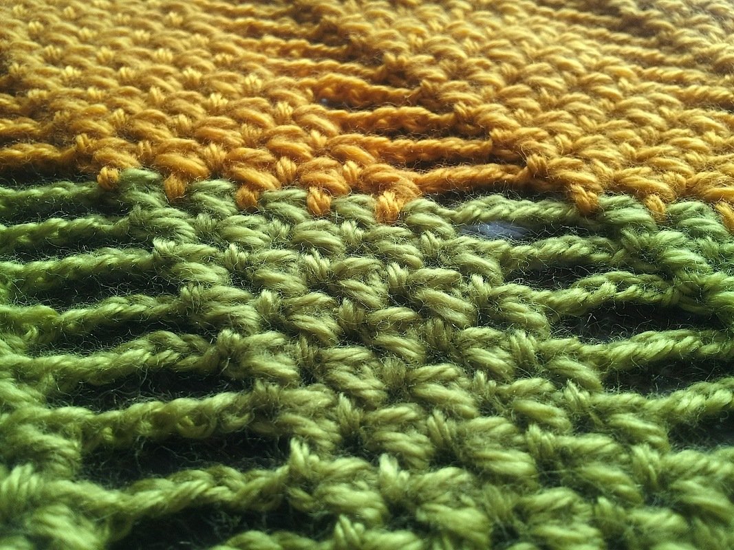 Close-up of crocheted fabric with golden-yellow yarn transitioning to green yarn, showing textured stitches.