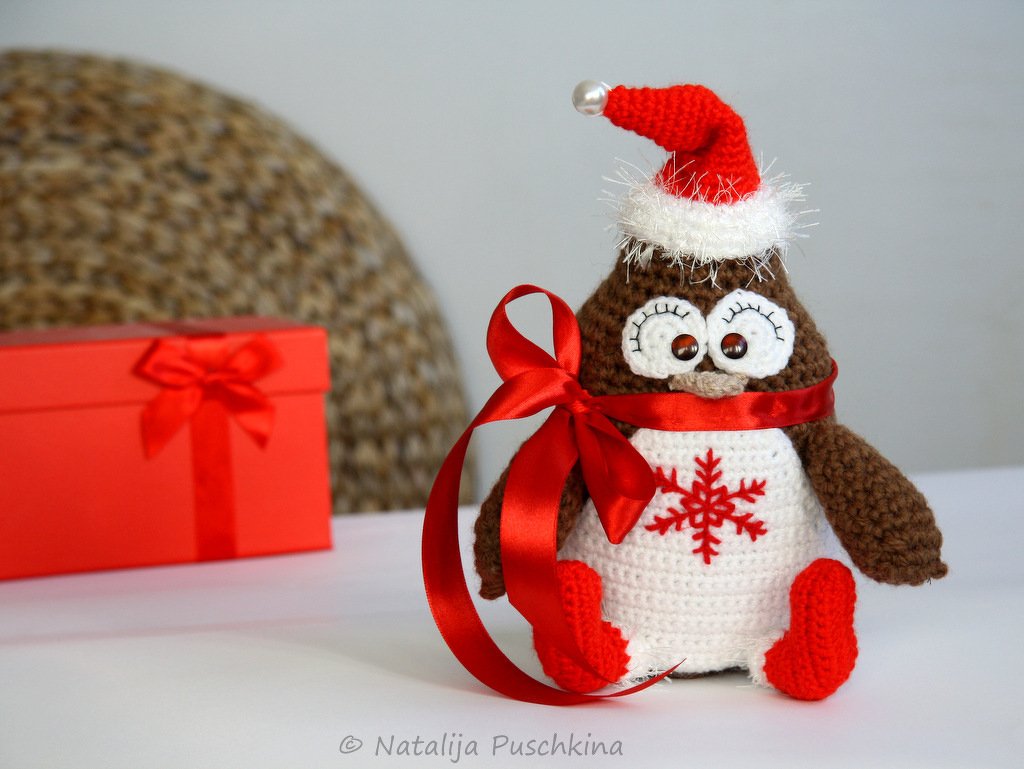 Gehäkelter Amigurumi-Pinguin mit roter Weihnachtsmütze, roter Schleife und Schneeflockenmotiv neben rotem Geschenk