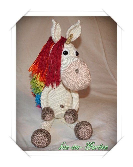 Sitzendes gehäkeltes Einhorn-Amigurumi mit regenbogenfarbener Mähne und goldfarbenem Horn.