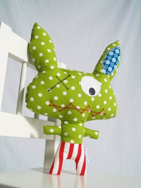 Grünes Punkte-Kuscheltier mit einem Auge, besticktem Mund, blau gemustertem Innenohr und rot-weiß gestreifter Hose.
