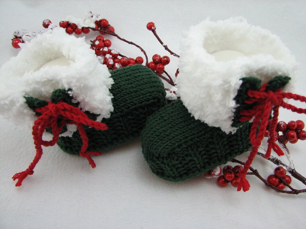 Paar gestrickter dunkelgrüner Babyschuhe mit weißem Umschlag und roten Schnürbändern vor roten Beeren