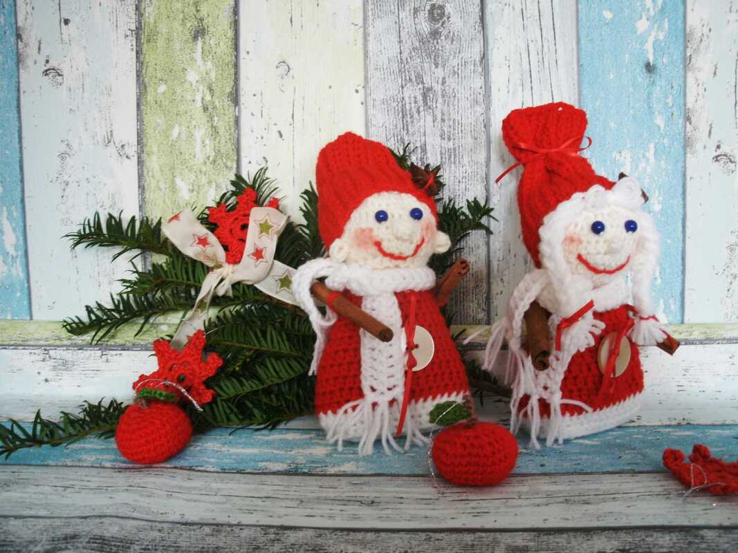 Zwei gehäkelte Weihnachtswichtel mit roten Mützen, blauen Knopfaugen und Zimtstangen vor rustikalem Holz mit Tannenzweigen und Häkeldekoration.