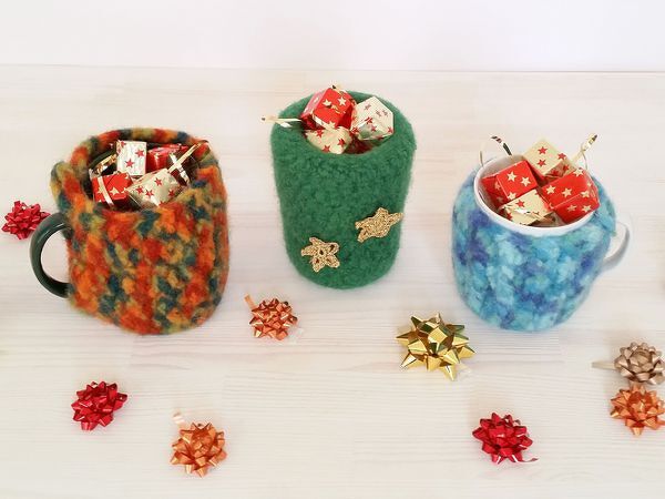 Reihe von drei Tassen mit gehäkelten, gefilzten Hüllen und einem gefilzten Behälter, gefüllt mit kleinen Geschenkwürfeln.