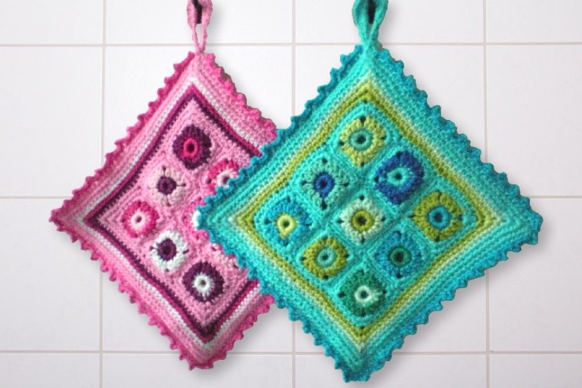 Zwei gehäkelte Granny-Square-Topflappen in Pink und Türkis hängen an einer gefliesten Wand.