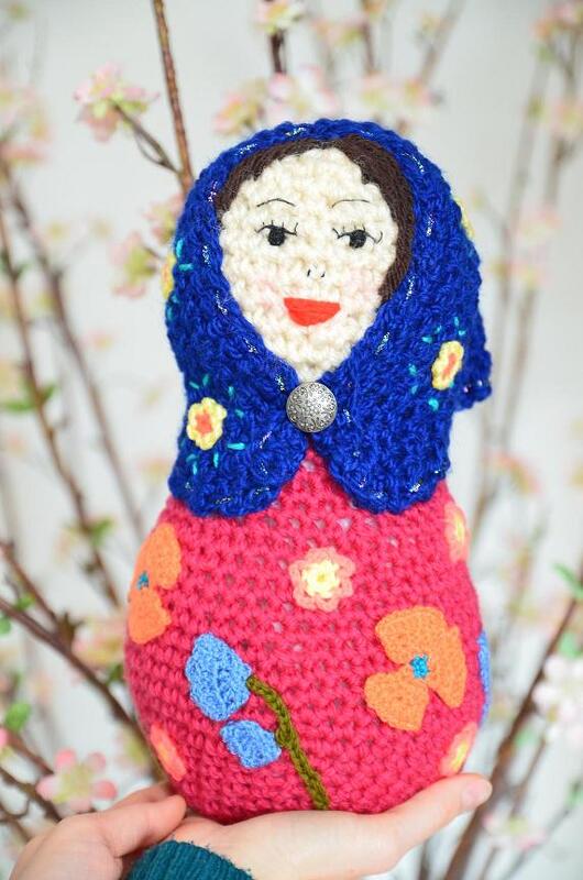 Gehäkelte Amigurumi-Matroschka mit blauem Kopftuch und pinkem Körper mit Blumenapplikationen