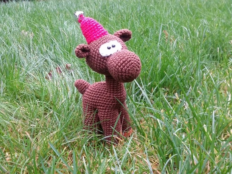 Gehäkeltes braunes Amigurumi-Rentier mit pinker Zipfelmütze und Glöckchen im Gras
