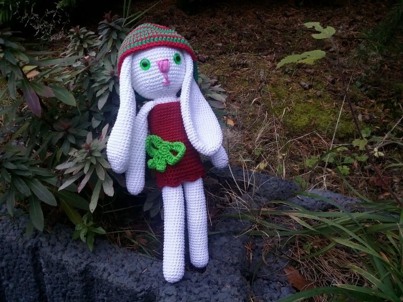 Amigurumi-Hase mit weißem Körper, rotem Kleid, grüner Schleife und gestreifter Mütze auf dunklen Steinen