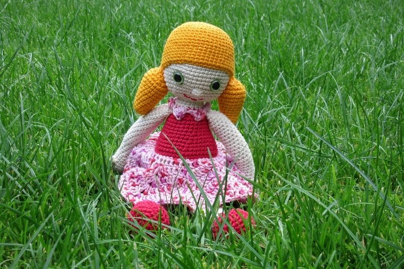 Gehäkelte Amigurumi-Puppe mit orangefarbenen Zöpfen und pink-rotem Kleid, die im grünen Gras sitzt