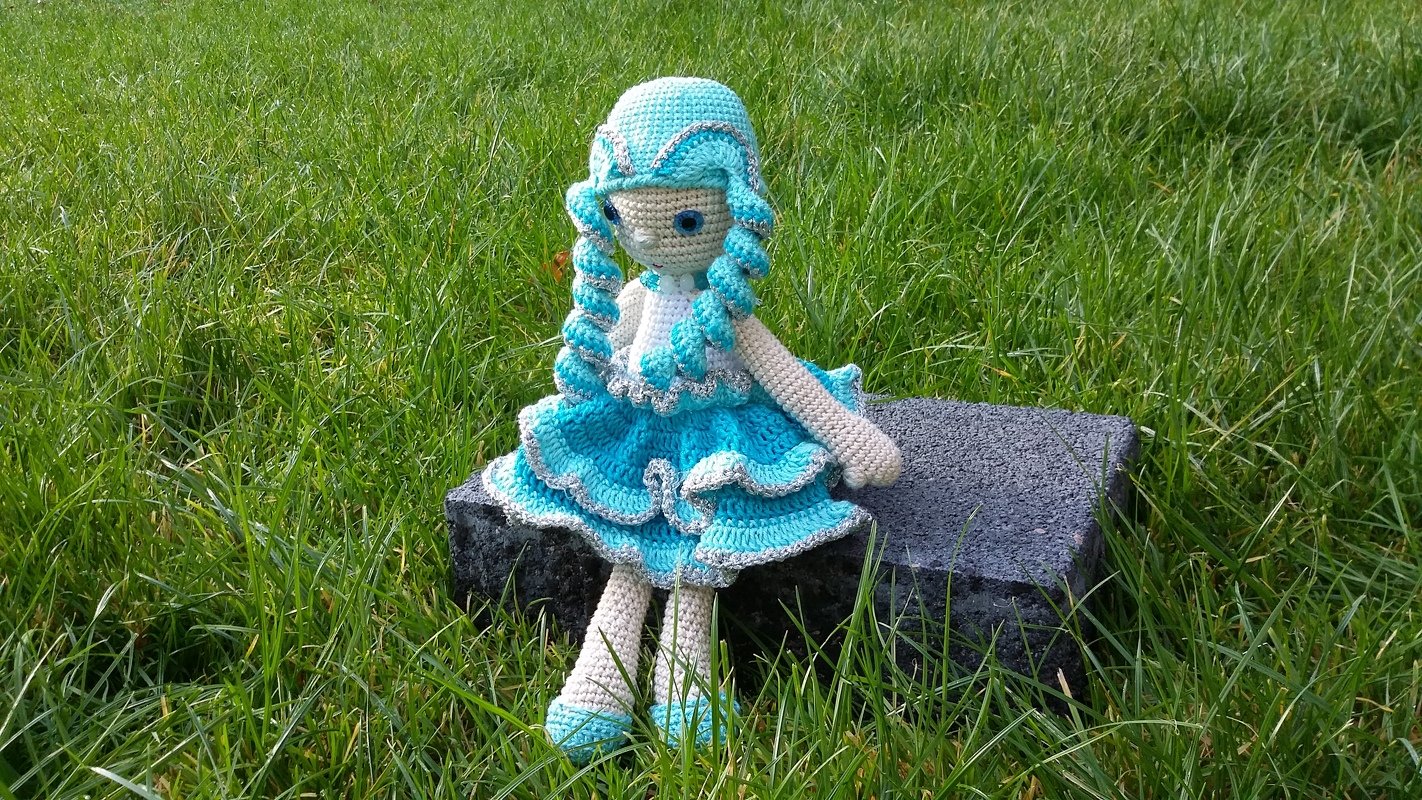 Gehäkelte Amigurumi-Puppe in türkis-weißem Rüschenkleid mit gedrehten Zöpfen, sitzend auf Stein im Gras