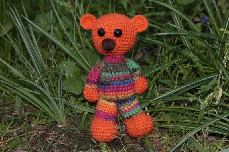 Gehäkelter Amigurumi-Teddybär mit orangem Kopf und bunt gestreiftem Körper zwischen grünen Pflanzen.