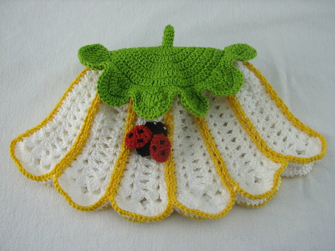 Gehäkelter Blütenhut in Weiß und Gelb mit grünem Blatt und zwei roten Marienkäfern auf hellem Hintergrund