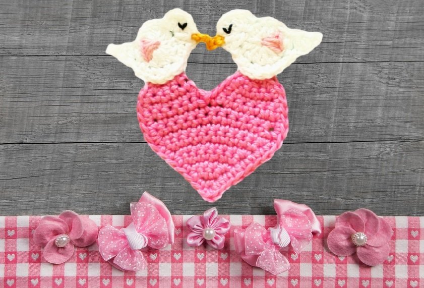 Zwei weiße gehäkelte Vögel mit gelben Schnäbeln berühren sich über einem rosa Häkelherz; darunter rosa Stoffblumen und Schleifen auf kariertem Stoff.