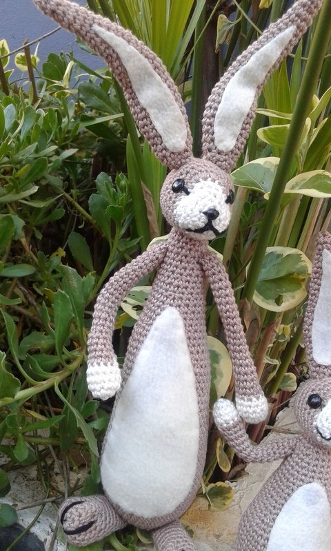Beige-weißer gehäkelter Amigurumi-Hase mit langen Ohren und weißem Filzbesatz vor grünen Pflanzen