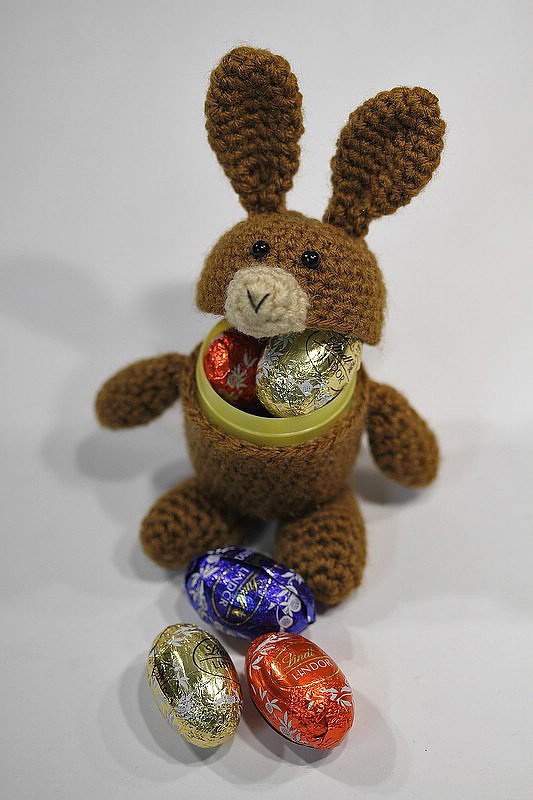 Brauner gehäkelter Amigurumi-Hase mit Öffnung am Bauch, darin bunt verpackte Schoko-Eier; drei Eier liegen davor.