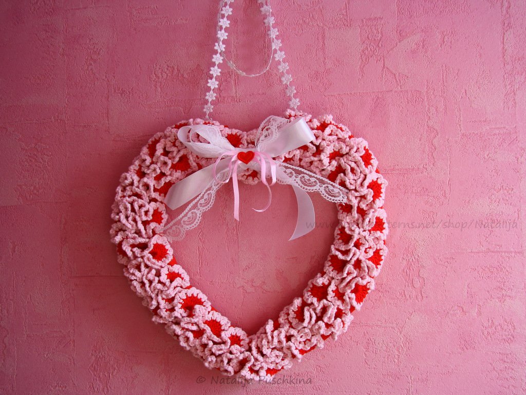 Gehäkeltes Herz aus rosa und roter Wolle mit weißer Schleife, Spitze und kleinem roten Herzknopf an einer rosa Wand.
