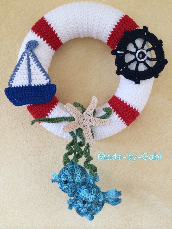 Gehäkelter Rettungsring-Kranz mit rot-weißen Streifen, blauem Segelboot, Steuerrad, Seestern und gehäkelten Fischen.
