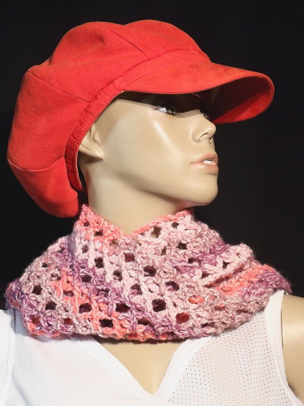 Schaufensterpuppe trägt gehäkelten Loop-Schal in Rosa, Lila und Orange sowie eine rote Kappe vor dunklem Hintergrund.