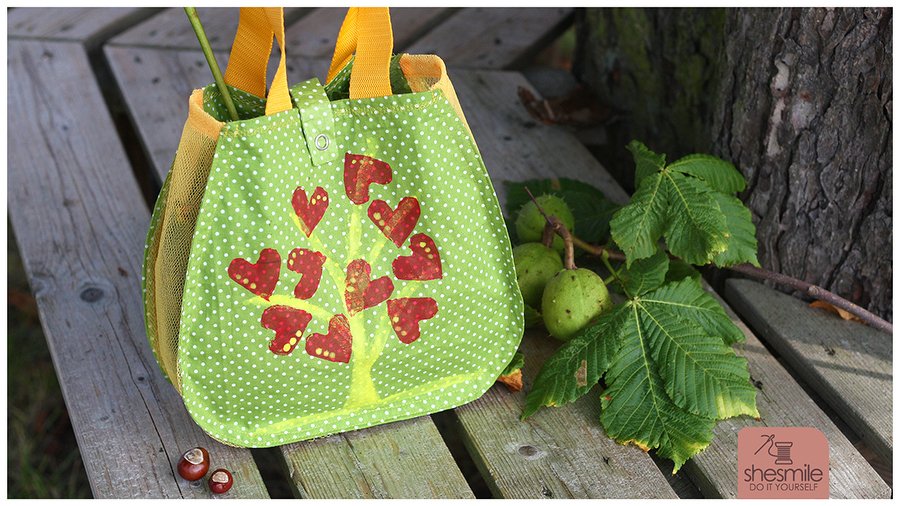 Grüner Pünktchen-Stoffbeutel mit Herzbaum-Motiv und gelben Henkeln auf Holzbank neben Kastanien