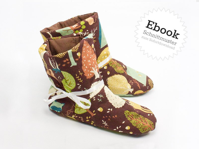 Braune Kinderhausschuhe mit buntem Wald- und Baummuster, weißes Band, vor hellem Hintergrund.