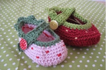 Zwei gehäkelte Babyschuhe; links rosa mit grünem Riemen und Punkten, rechts rot mit grünem Rand und Knopf