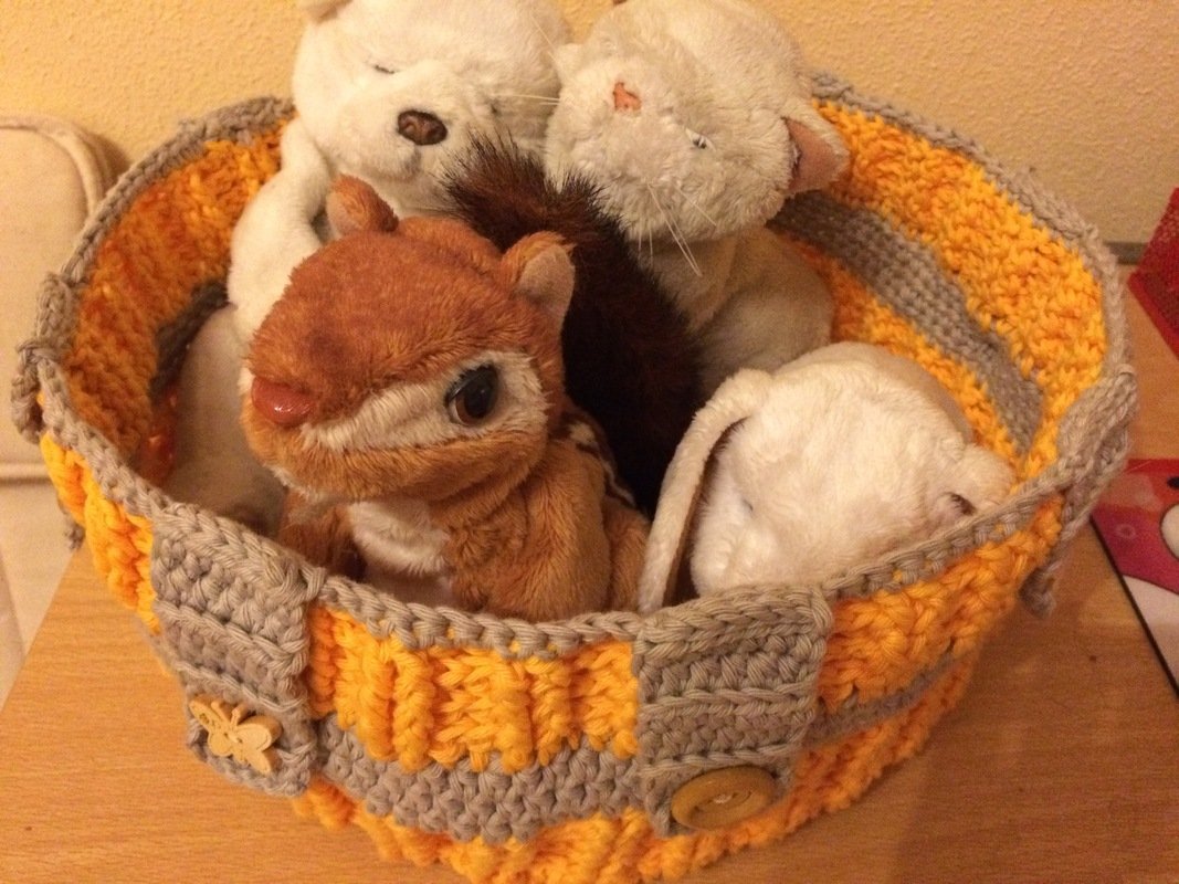 Gehäkelter Utensilo-Korb in Gelb, Orange und Grau, gefüllt mit Plüschtieren und Stofftieren.