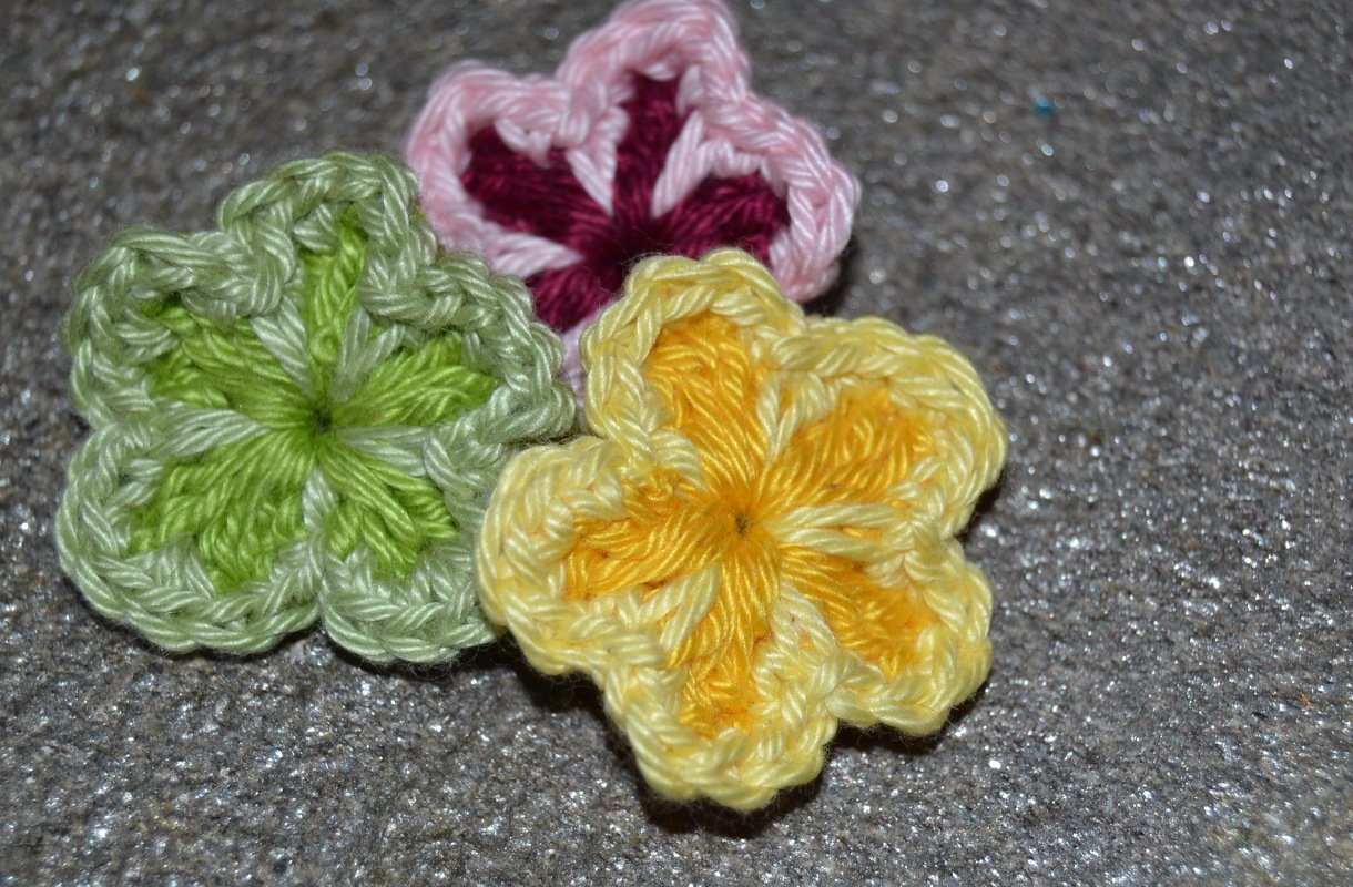 Drei gehäkelte Blumenapplikationen in Gelb, Grün und Rosa auf glitzerndem grauem Untergrund