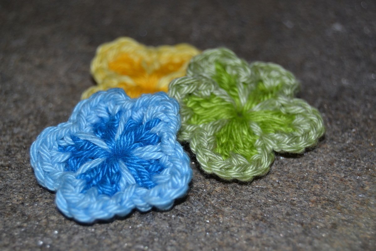 Drei gehäkelte Blumen‑Applikationen in Blau, Grün und Gelb auf dunklem Hintergrund.