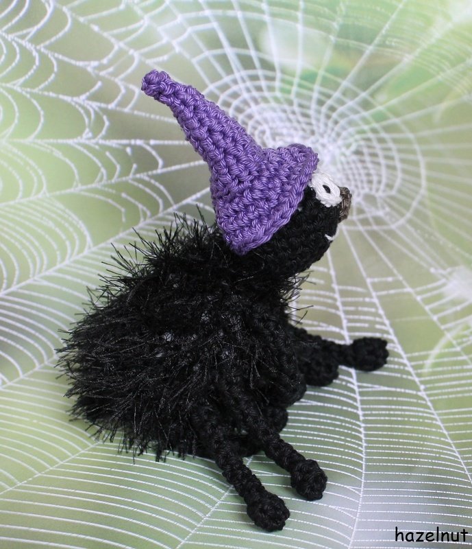 Gehäkelte schwarze Amigurumi-Spinne mit lila Hexenhut vor Spinnennetz-Hintergrund