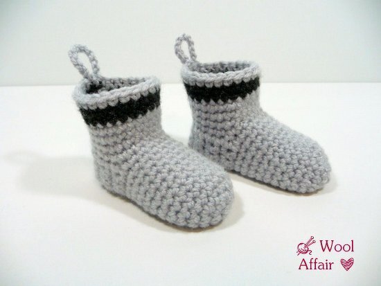Zwei hellgraue, handgehäkelte Babysocken mit schwarzem Streifen am Schaft und kleinen Schlaufen an den Fersen vor neutralem Hintergrund.