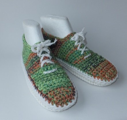 Gehäkelte Sneaker-Hausschuhe, grün-braun meliert, mit weißen Schnürsenkeln und weißer Sohle auf hellem Hintergrund.