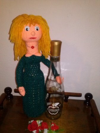 Gehäkelte Puppe mit blonden Haaren und grünem Kleid steht neben einer Flasche auf einem Holztisch, davor rote Blüten.